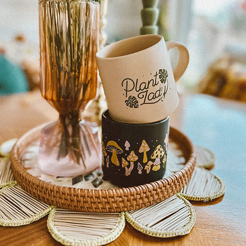 Two mugs are stacked on top of a bohemian table setting. One is tan and features hand lettering reading Plant Lady with monstera leaf illustrations in green ink. The second is black with pink and yellow mushroom illustrations wrapping around the mug.