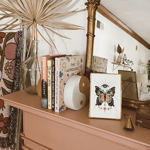 An art print of a hand illustrated butterfly with all knowing eye and stars sits atop a terra cotta painted mantle with books, ritual tools, decorative vases and a beautiful ornate mirror.
