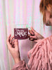 A bohemian woman in a pink shag sweater holds a clear glass mug with hand lettering that says "You are magic" in front of a sparkling irridescent backdrop. The mug has whimsical clouds and twinkling stars surrounding the beautiful hand lettering.