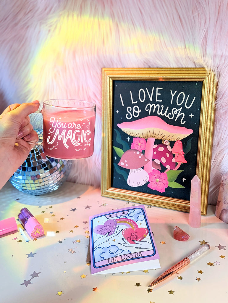 Someone holds a glass mug in front of a bohemian setting featuring beautiful mushroom illustrations, tarot cards, disco ball, crystals, star confetti, sparkling pens, and Trixie Cosmetics lipgloss. The mug says You are magic in a playful hand lettering with whimsical clouds and magical twinkling stars around it.