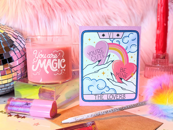 A Valentine's Day Card is shown next to a feather pen, hand lettering mug, Trixie Cosmetics lipgloss and vibrant discoball. The card features artwork inspired by The Lovers tarot card. There are two hands each holding a candy heart with hand lettering that says "You're Cute" and "Be Mine" A rainbow connects the hearts and in the background there are whimsical clouds and twinkling stars. A truly magical card to celebrate your love.