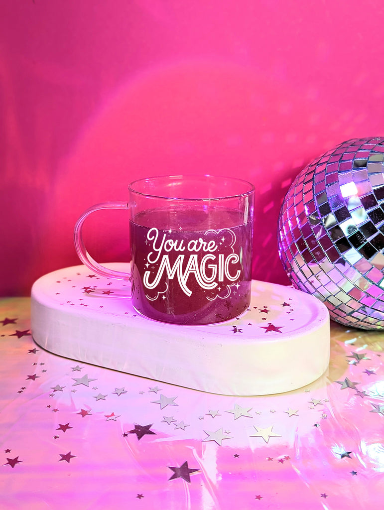 A glass mug sits in front of a discoball and pink background surrounded by twinkling star confetti. The Mug says "You are magic" in bold and playful hand lettering, surrounded by whimsical clouds and magical stars.