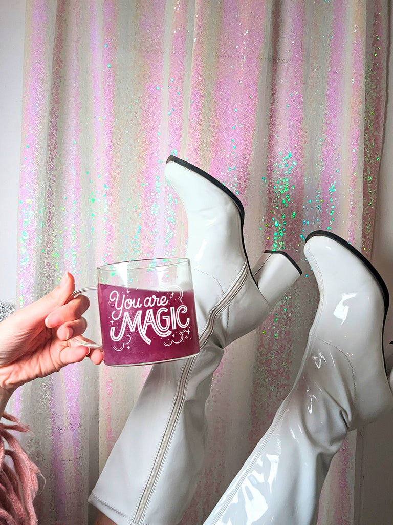 A woman holds a mug while kicking up her go go boots in front of a sparkling iridescent backdrop. The mug says You are Magic in playful and bold hand lettering, with whimsical clouds and twinkling magical stars. 