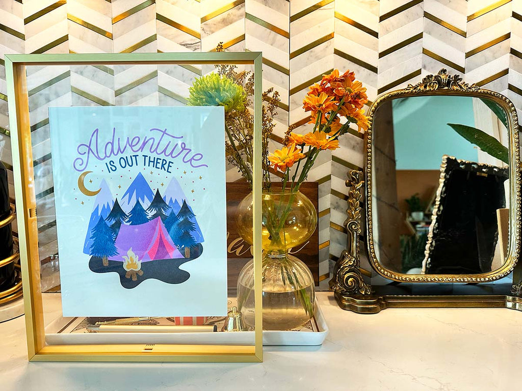 An art print in a modern gold float glass frame sits in front of a bohemian interior space. The illustration is of a cozy camping scene with a pink tent and roaring campfire tucked into a luscious whimsical forest scene with mountains and a starry sky in the background illuminated by the moon. Hand lettering spells out "Adventure is out there"