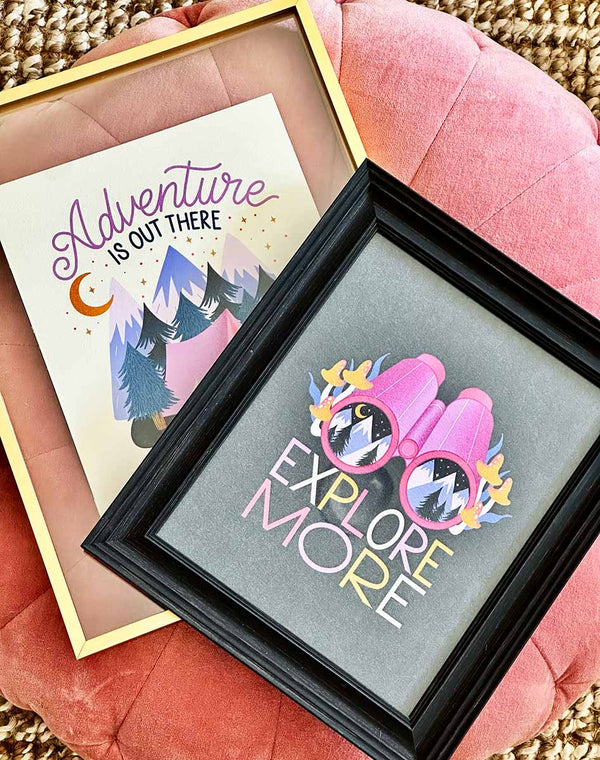 Two framed art prints sit atop a pink pillow pouf. The top is of a set of binoculars with a mountain scene inside. Mushrooms and squiggly grass frame the scene and hand lettered below is "Explore More". Behind it is another art print, with an illustration of a vibrant pink tent nestled in a forest scene with a roaring campfire. In the background are whimsical mountains, and a crescent moon illuminating the twinkling starry sky. Hand lettering spells out "Adventure is out there"