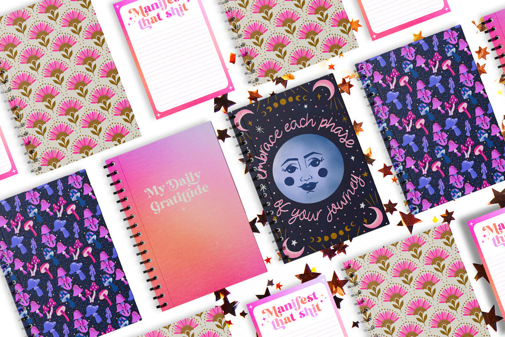 A collection of vibrant, colorful notebooks and notepads are on a white background with gold star confetti all around. The notebooks have a whimsical mushroom pattern, modern floral pattern, and moon and stars illustration that says "Embrace Each Phase of Your Journey". There is a guided gratitude journal with a pink aura background that says "My daily gratitude" in a retro inspired lettering style, and a cute notepad with pink ombre background that says "manifest that shit" in a pink and purple ombre.