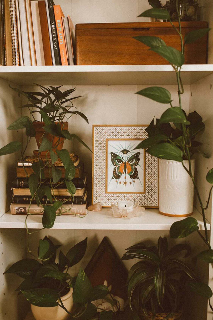 An art print in a moroccan frame sits atop shelves in a trendy bohemian interior. It is surrounded by candles, crystals, books, and luscious houseplants. The illustration features a butterfly with moon phases and cosmic eyes in its wings crying golden tears that pool into bright stars at the bottom. There is a star filled eye at the top and light beams emanating from it.