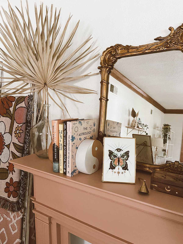 A framed art print in a trendy bohemian space atop a pink mantel. There are floral patterned blankets, dried palm in glass vases, astrological books and a yin yang book end all in front of a gorgeous gold framed mirror. The illustration is of a pink teal and black butterfly with moon phases in its bottom wings. In the top wings are a pair of intuitive eyes, filled with stars, and crying golden tears. Above the butterfly is an eye with a star inside, and below are gold stars. 