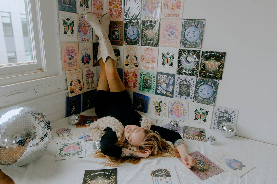 Photo is of Michigan based illustrator Kelsey DeLange surrounded by her collection of vibrant, uplifting art prints. Some of the designs feature cosmic themes, the moon, butterflies, bookish themes, whimsical mushrooms, and uplifting hand lettered mantras. There are discoballs scattered throughout