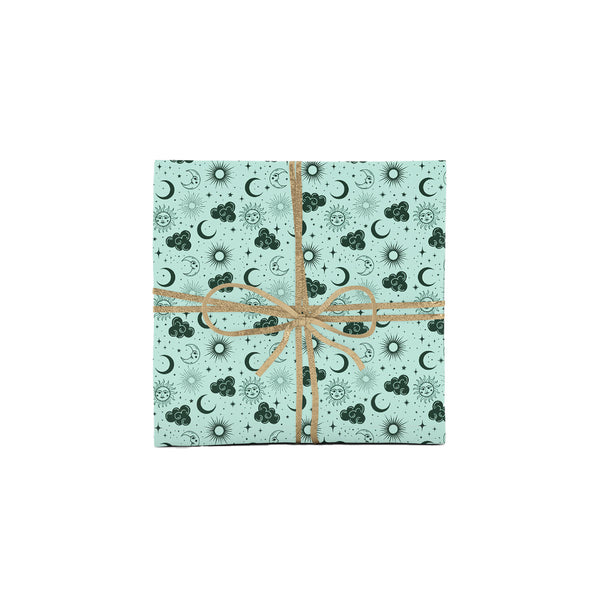 Photo shows a gift wrapped on a white background. The gift is wrapped in hand illustrated bohemian wrapping paper with a magical pattern of celestial symbols: suns, moons, clouds, and twinkling stars. The gift is wrapped in a gold bow.
