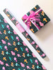 Photo shows a sheet of whimsical hand illustrated wrapping paper , roll of the wrapping paper, and a gift, wrapped with a lovely pink ribbon. The wrapping paper has patterns of colorful mushrooms surrounded by greenery and twinkling stars.
