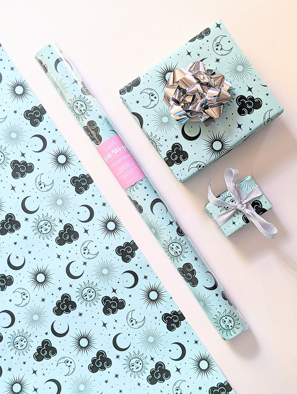 Photo shows a sheet of wrapping paper, roll of gift wrap and two gifts wrapped with silver bows. The wrapping paper has a bohemian hand illustrated magical pattern of celestial symbols like suns, moons, clouds, and twinkling stars.
