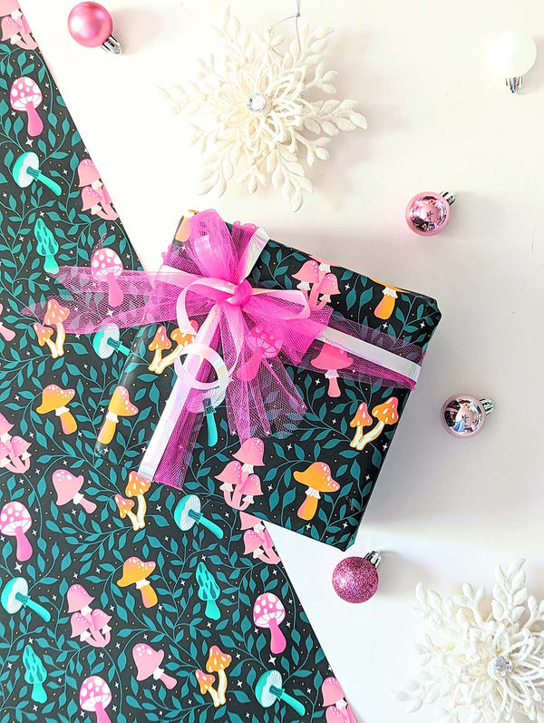 Photo shows a sheet of whimsical hand illustrated wrapping paper and a gift, wrapped with a lovely pink ribbon. The wrapping paper has patterns of colorful mushrooms surrounded by greenery and twinkling stars.