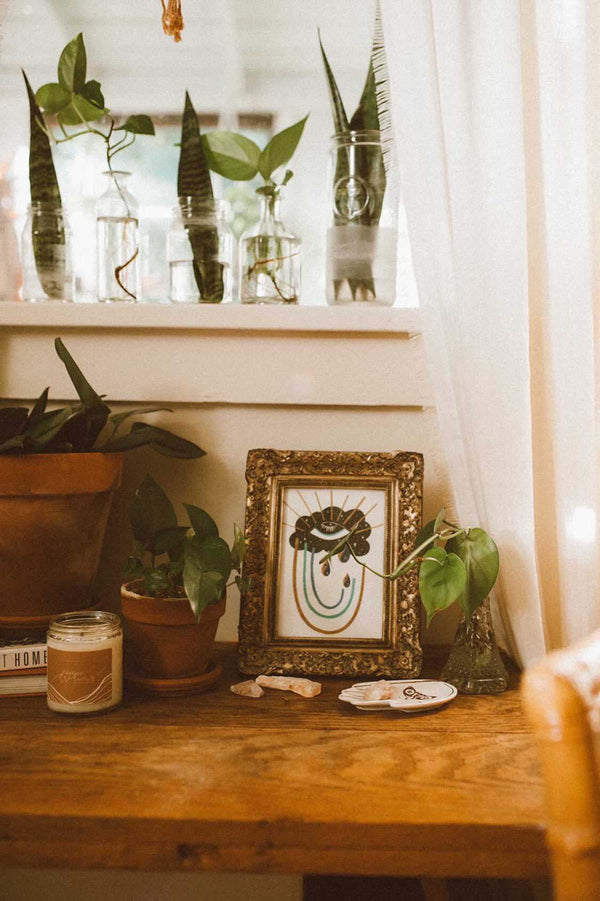 A lovely art print is framed in an ornate gold frame in a bohemian interior with house plants, candles, and crystals all around. A framed art print - the illustration is of a cosmic intuitive eye in a starry cloud, it sheds tears that are small yin yangs, the eyelashes as rays emanate from above and a rainbow of vibrant blues and golds emerge at the bottom. Signifying the beauty that comes with clarity.