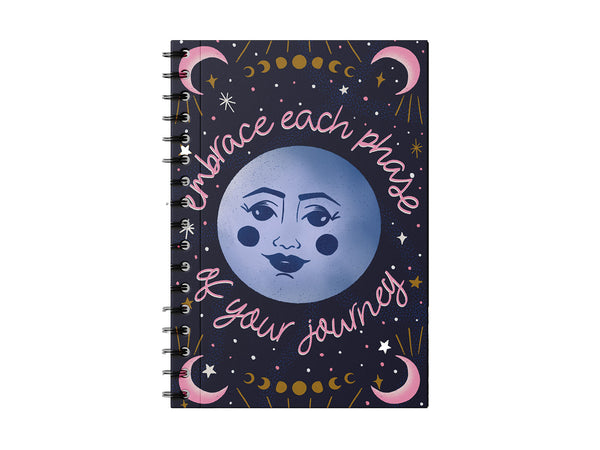 Photo shows a notebook on a white background. The notebook has a dark blue cover with a full moon in the center. Around it is hand lettered in pink script "Embrace each phase of your journey", there are crescent moons in the corners, and all of the moon phases illustrated and there are stars and twinkles all throughout the design. It's a lovely, cosmic bohemian notebook.