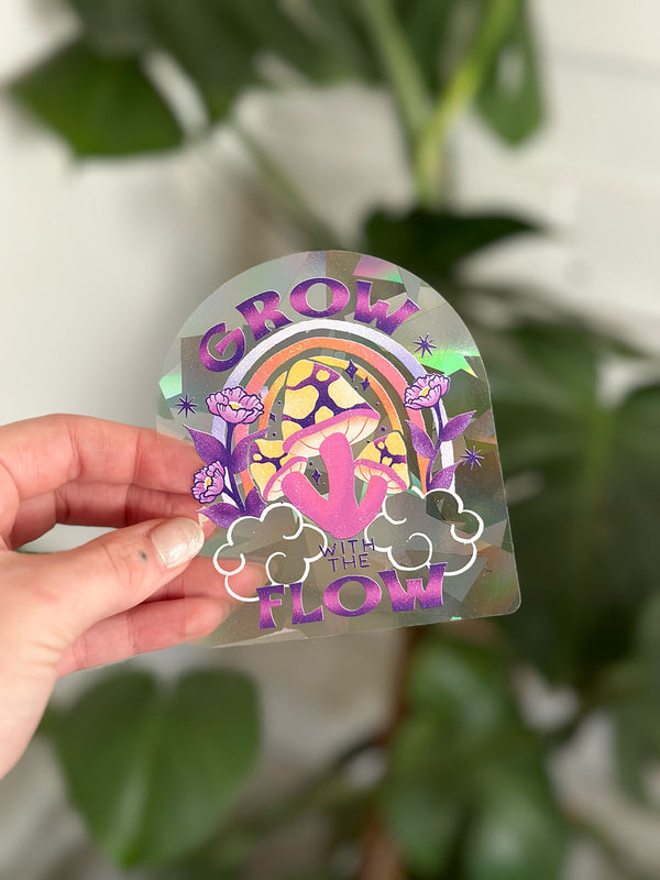 a rainbow maker sun catcher sticker is held in front of a vibrant monstera plant. The sticker reflects the sun into rainbows all over your home. It has an illustration of colorful mushrooms, flowers, and a rainbow in the background and says "grow with the flow" in a playful retro inspired hand lettering style. Perfect for bohemian decor.
