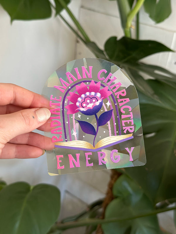 Photo shows a rainbow maker sun catcher sticker in front of a vibrant monstera plant. The sticker goes on the window and reflects sunlight into rainbows all over your home. It has an illustration of an open book with a vibrant flower blooming from its pages. There is a rainbow in the background. It says "Radiate Main Character Energy" in a playful, retro inspired hand lettering style. Perfect gift for a bookworm, reader, or booktok lover and great addition to your bohemian decor.  