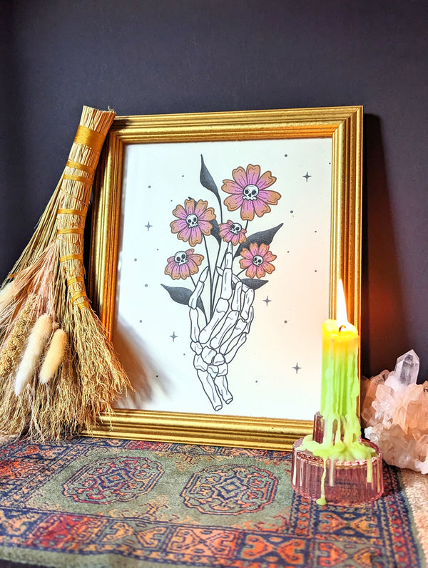 A witchy art print is framed beside a glowing green candle, crystals, and dried florals. A framed art print - the illustration is of a skeleton hand holding a bouquet of vibrant orange and purple flowers that have small skull heads in the center. It is surrounded by little twinkles for a fun, whimsical addition to your bohemian Halloween decor.