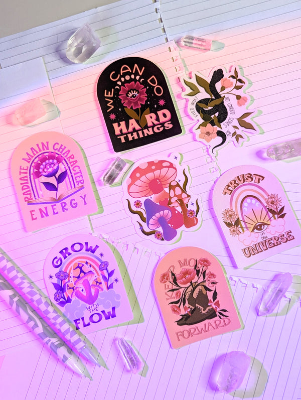 A collection of hand illustrated stickers sits atop notebook pages surrounded by clear quartz crystals. There are astrological stickers for each zodiac sign. Aries, Taurus, Cancer, Gemini, Leo, Virgo, Libra, Scorpio, Sagittarius, Capricorn, Aquarius, Pisces. There are Mushroom stickers, snake stickers, flower stickers, rainbow stickers. All with uplifting hand lettering messages. Witchy. Witch. Boho. Magical. Bohemian. Yoga. Yogi.
