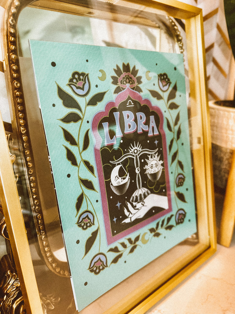 A framed art print is in a gorgeous gold float glass frame. A framed art print - the illustration is a depiction of the Libra zodiac sign. The celestial scale is illustrated floating in a starry night sky, surrounded by whimsical clouds and framed in by folk art flowers. Libra is hand lettered at the top in a bold, groovy, retro inspired style.
