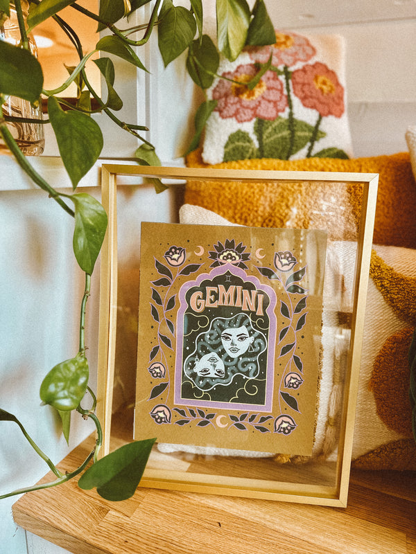 A framed art print sits in a bright bohemian interior space surrounded by trendy pillows and luscious house plants. A framed art print - the illustration is a depiction of the Gemini zodiac sign. The two twins are floating in a celestial sky with whimsical moons and twinkling stars all around. Framed by folk art inspired florals with Gemini hand lettered at the top in a groovy, retro inspired style.