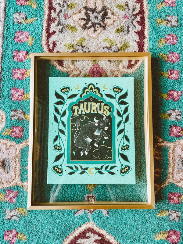 A framed art print sitting atop a bold, vibrant, bohemian style moroccan rug. A framed art print - the illustration is a depiction of the Taurus zodiac sign. The celestial bull is illustrated floating in a starry night sky, surrounded by whimsical clouds and framed in by folk art flowers. Taurus is hand lettered at the top in a bold, groovy, retro inspired style.