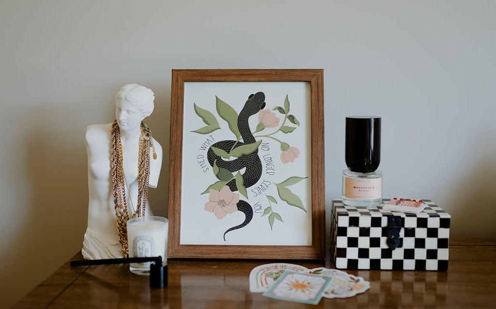 A framed art print sits in a modern, contemporary interior space with a statue, candles, and checkered box surrounding it. A framed art print - the illustration is of a black snake coiled and slithering through vibrant pink florals and greenery. Hand lettering spells out "Shed what no longer serves you" on either side.