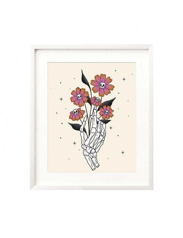 A framed art print - the illustration is of a skeleton hand holding a bouquet of vibrant orange and purple flowers that have small skull heads in the center. It is surrounded by little twinkles for a fun, whimsical addition to your Halloween decor.