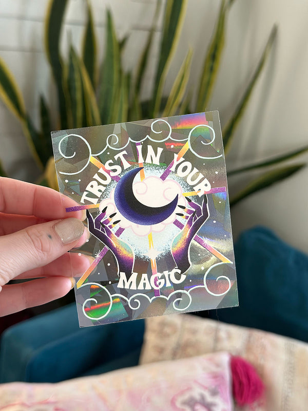 Photo shows a rainbow maker sun catcher sticker in front of a vibrant snake plant. The sticker goes on the window and reflects sunlight into rainbows all over your home. It has an illustration of two magical rainbow hands summoning the power of the moon, surrounded by dreamy clouds and twinkling stars. It says “Trust in Your magic” in a playful, retro inspired hand lettering style. Perfect gift for a witchy, intentional magic woman and great addition to your bohemian decor.  