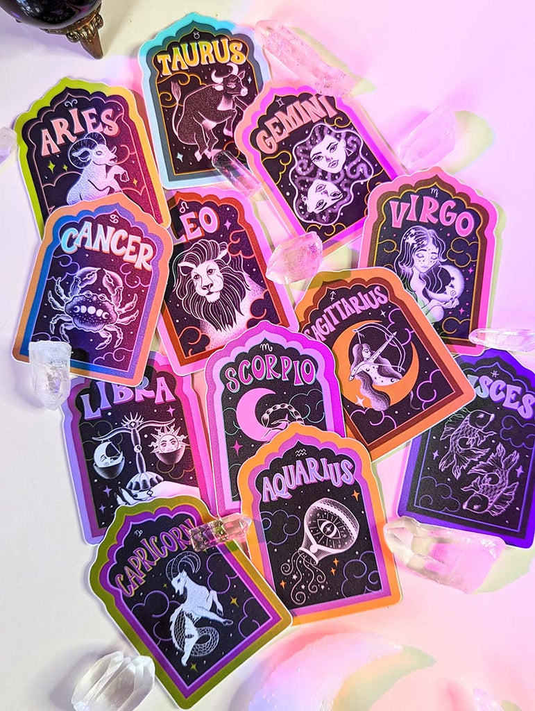 A collection of zodiac stickers sits on a white background with purple aura glow lights. There are illustrated stickers of each astrological sign, with a retro hand lettering spelling out the name of each sign. Stickers for Aries, Taurus, Cancer, Gemini, Leo, Virgo, Libra, Scorpio, Sagittarius, Capricorn, Aquarius, and Pisces. Water sign, Fire Sign, Earth Sign, Air Sign. Surrounded by clear quartz crystals.