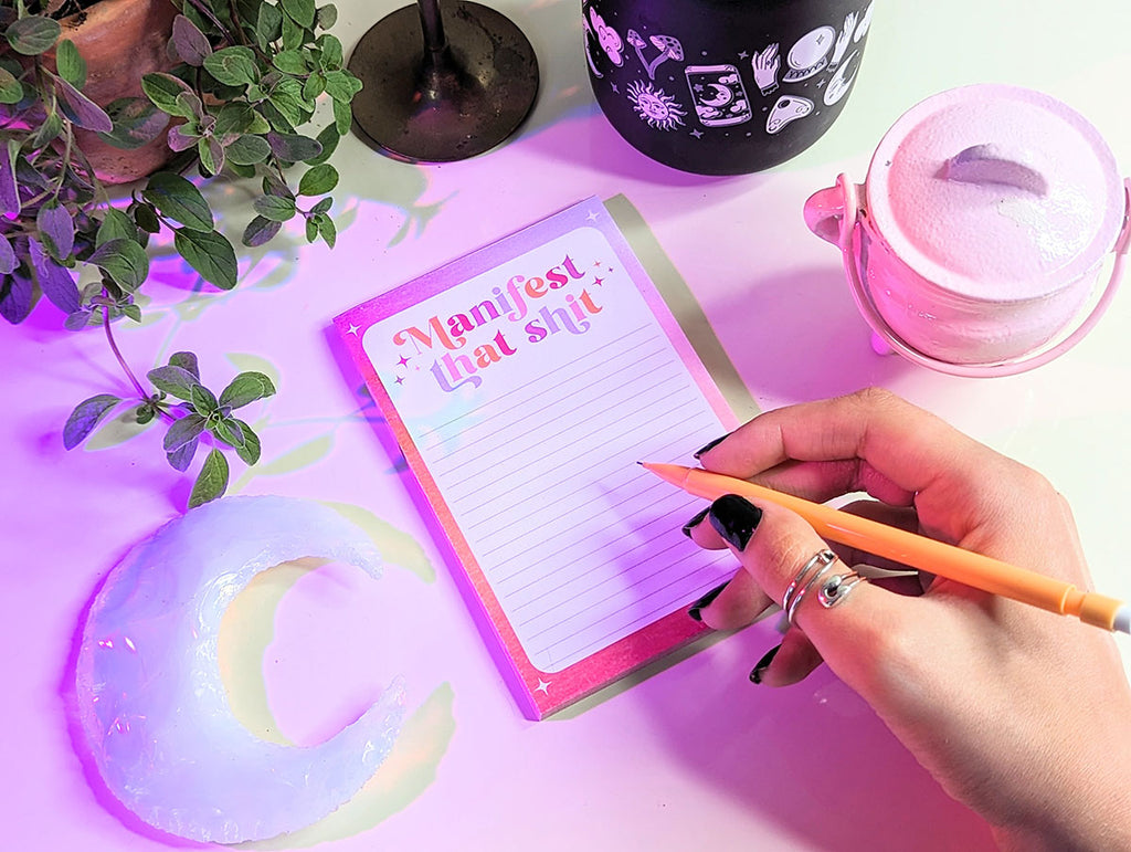 A notepad sits on a white desk with pink and purple aura lighting. A hand with black nails and boho rings hold a pencil above a magical notepad that says "Manifest that Shit" in a retro inspired lettering style. It has illustrated stars around it. There are plants and crystals and a mug in the background. This notepad is perfect for keeping track of your goals, accountability, mindfulness, meditation, and achieving your goals.