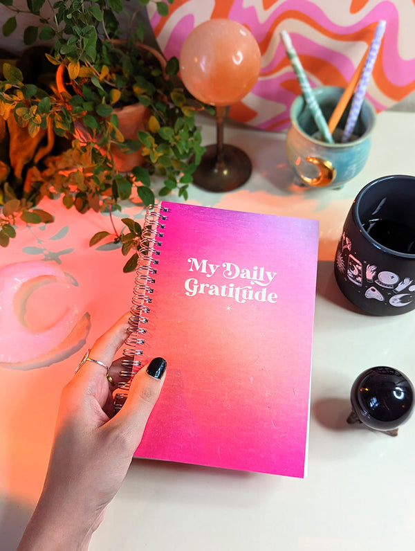 A hand with black painted nails holds a vibrant pink aura journal that says "My Daily Gratitude" on the cover. It is a mindfulness journal to reflect and meditate on what you're grateful for each day. There is a funky, vibrant, retro desk scene in the background with a magical mug, crystal ball, crescent moon shaped crystal, and mystical mug. colorful bohemian decor.