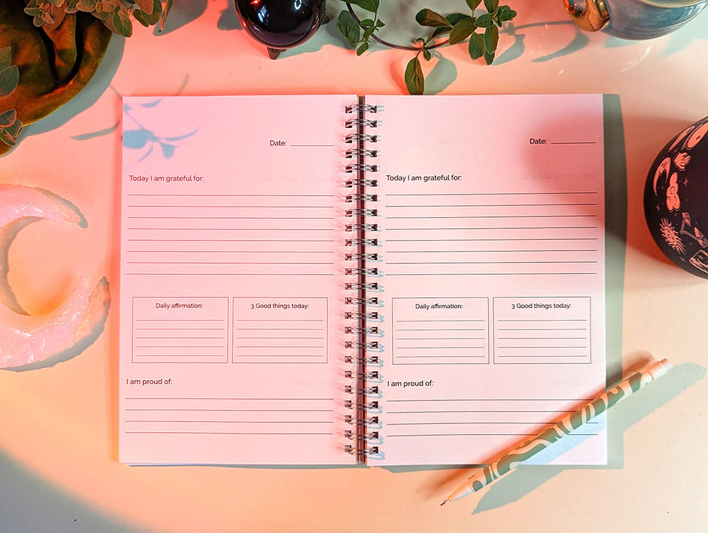 A guided gratitude journal sits open on a white background. The prompts include "today i am grateful for", "daily affirmation", "3 good things today", and "I am proud of" with space to write and reflect for each. Lovely mindfulness tool for yoga, bohemian, magical mindful stationery. There are crescent moon shaped crystals plants and a psychedelic design pencil.