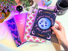 A pile of notebooks sit on a white desk with glowy purple pink aura lighting. They are surrounded by crystals and house plants. The top one has an illustration of the moon and hand lettering says "Embrace each phase of your journey". Behind it is a pink gradient cover that says "My Daily Gratitude" this is a guided journal to help you reflect and meditate on what you are grateful for each day. One has illustrated flowers on the cover in an art deco style the last one has whimsical mushroom illustrations.
