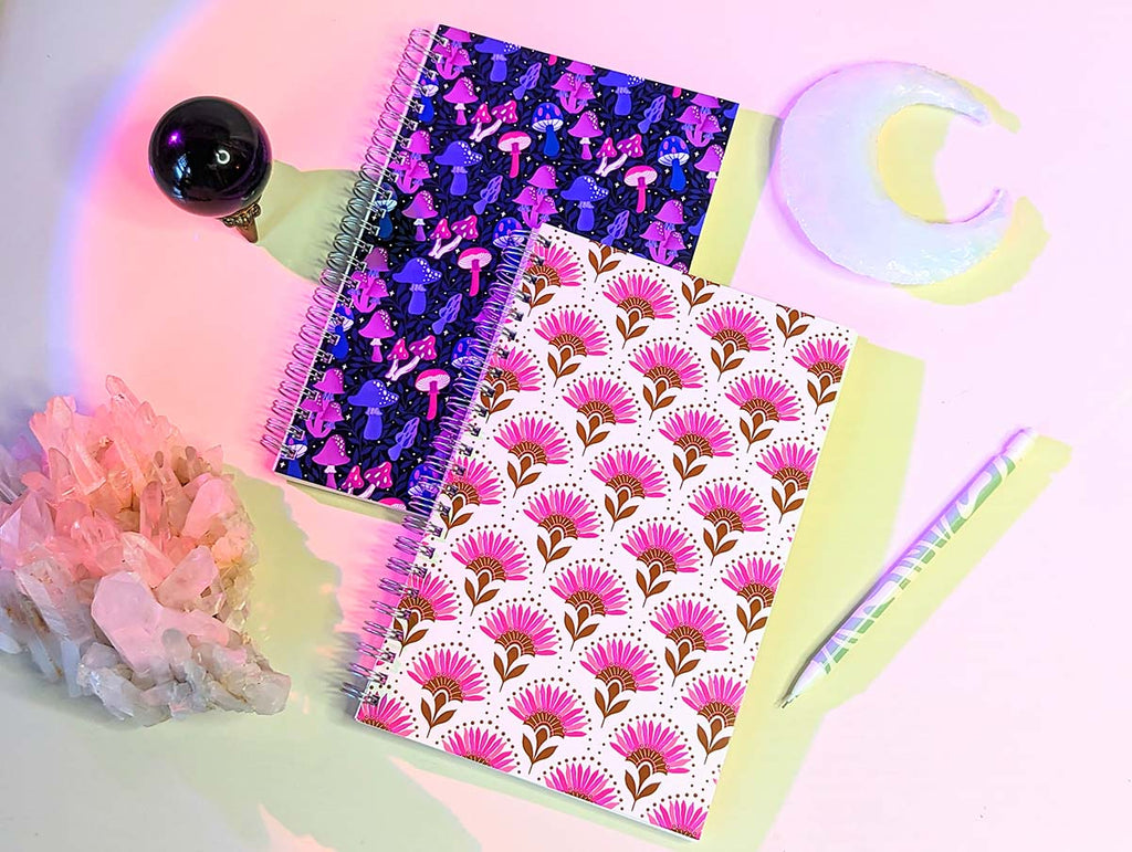 Hand illustrated notebooks sit on a white desk with pink aura lighting surrounded by crystal clusters and crescent moon shaped quartz. The top notebook has a pink and gold floral pattern in an art deco inspired style. The bottom notebook has a whimsical colorful mushroom pattern. They are perfect additions to your office or companions along your mindfulness and magical journey. Great gifts for the witchy, magical, bohemian, boho, retro, nature loving girl in your life.