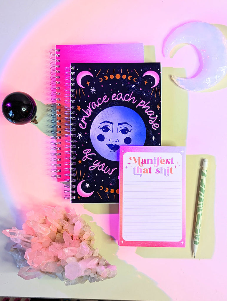 A pile of notebooks and notepads sits on a white desk surrounded by crystals, crescent moon quartz. The notepad says "Manifest that Shit" in a fun retro inspired lettering style in shades of pink, purple, and orange. The notebook has a moon illustration and says "embrace each phase of your journey" in a fun hand lettered style. They are perfect additions to your office decor or desk accessories and make great gifts for the boho, witchy, magical, retro, yogi, bohemian, ladies in your life.