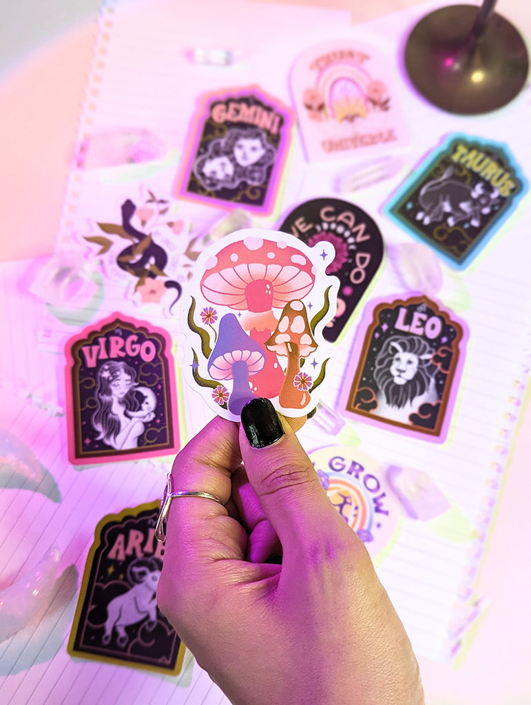 A witchy boho manicured hand holds an illustrated waterproof sticker with a whimsical psychedelic mushroom illustration in shades of pink purple and orange with daisies around. Behind it is a collection of other stickers surrounded by clear quartz crystals. There are zodiac stickers for each astrological sign. Aries, Taurus, Cancer, Gemini, Leo, Virgo, Libra, Scorpio, Sagittarius, Capricorn, Aquarius and Pisces. There are mushroom stickers, snake stickers, rainbow stickers, yoga stickers. 