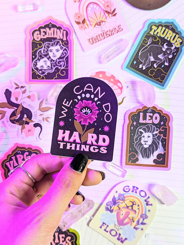 A witchy boho manicured hand holds an illustrated waterproof sticker with a flower that says "We Can Do Hard Things" in a retro hand lettering style. Behind it is a collection of other stickers surrounded by clear quartz crystals. There are zodiac stickers for each astrological sign. Aries, Taurus, Cancer, Gemini, Leo, Virgo, Libra, Scorpio, Sagittarius, Capricorn, Aquarius and Pisces. There are mushroom stickers, snake stickers, rainbow stickers, yoga stickers. All with uplifting messages and mantras.