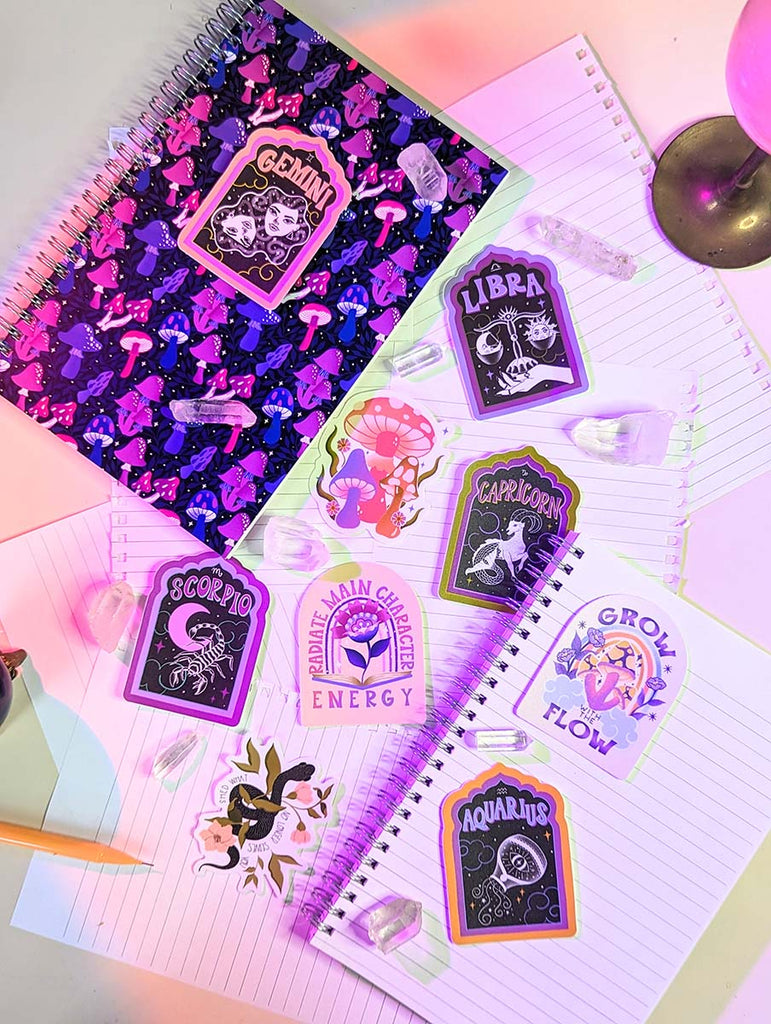 A notebook with hand illustrated mushrooms in various shades of pink, purple, and blue sits on a desk surrounded by crystals and stickers. The stickers have mushroom illustrations, zodiac illustrations, all astrological signs, libra, capricorn, scorpio, and aquarious, and a snake illustration. The stickers have uplifting mantras in retro inspired hand lettering styles. Perfect for the magical, witchy, boho, zodiac girlie, nature lover, yogi in your life.