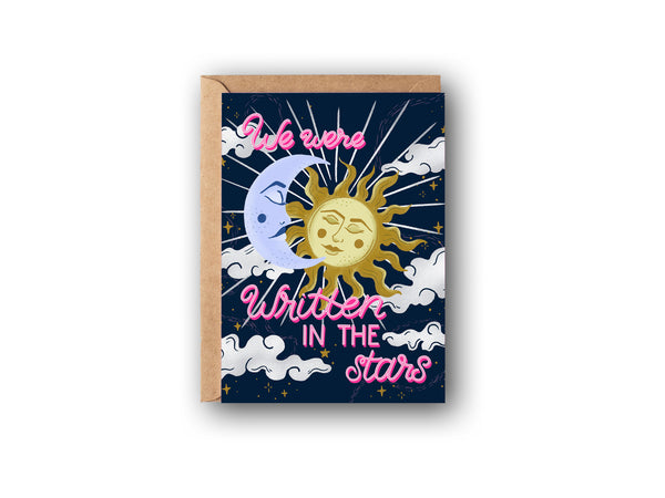 A Valentine's Day card is shown on a white background. The card features hand lettering spelling out "We Were Written in the Stars", there is an illustration of a celestial crescent moon and sun embracing and illuminating the night sky. There are whimsical clouds, twinkling stars, and magical star dust in the background. The perfect card to celebrate your love.