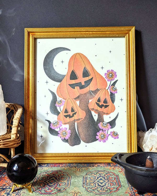 A witchy art print sits in a contemporary bohemian interior with incense burning in a cauldron pot, a small crystal ball, and moroccan rug. A framed art print - the illustration is of whimsical mushrooms drawn with jack o lantern faces, and colored orange for Halloween. It is surrounded by vibrant florals that have skull heads in the center and a crescent moon and twinkling stars make up the background. 