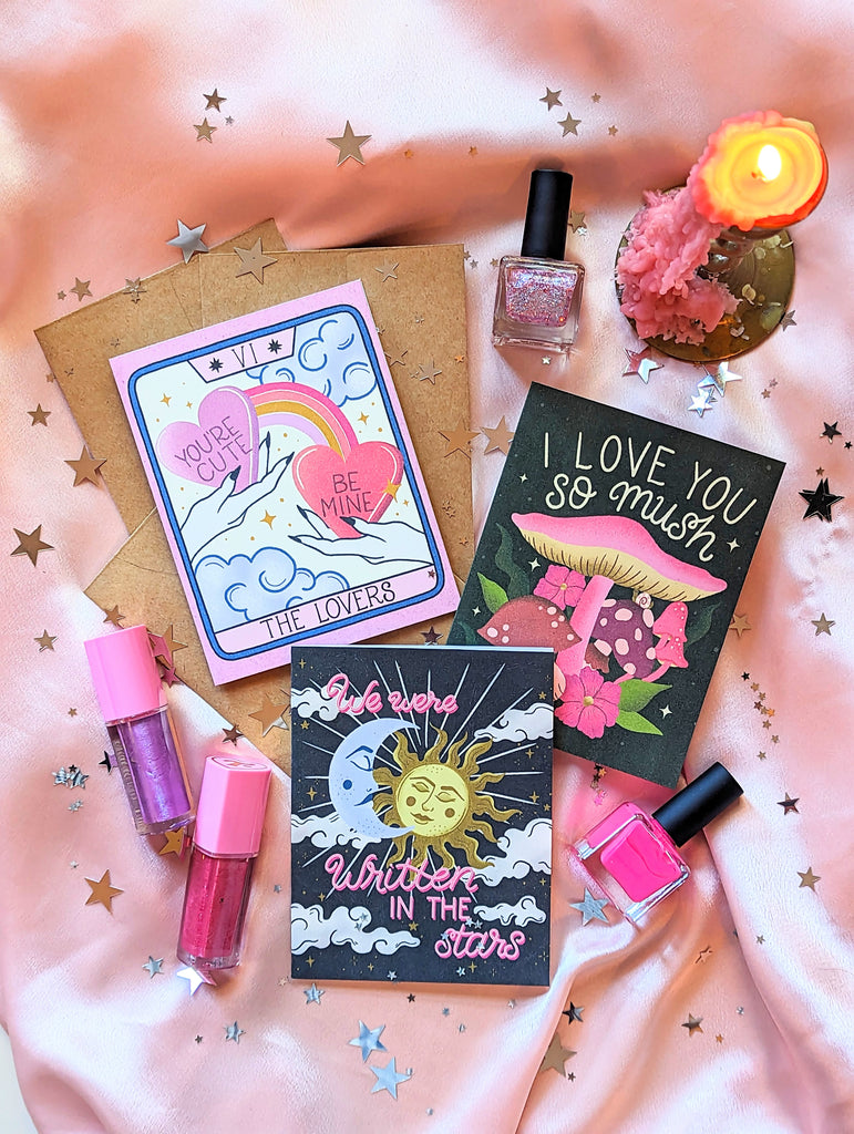 Three Valentine's Day cards are shown on a pink background with star confetti scattered about and Trixie Cosmetics lipgloss. The first card says "We Were Written in the Stars" in hand lettering script with a beautiful celestial crescent moon and sun illustration. The second is The Lovers Tarot Card, showing two hands with candy hearts and rainbows connecting them. Finally the last one says "I Love you so Mush" in hand lettering with vibrant mushrooms and fungi.