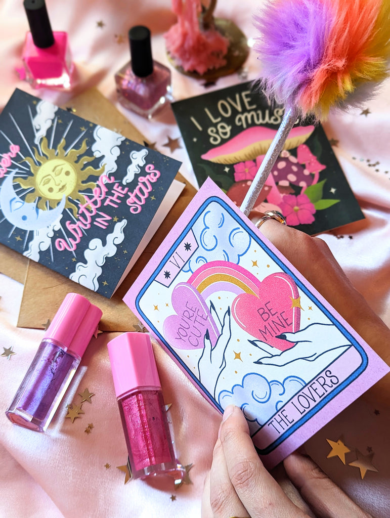 Three Valentine's Day cards sit on a pink background surrounded by star confetti and Trixie Cosmetics lip gloss. In the background one says "We Were Written in the Stars" in hand lettering with illustrations of a celestial sun and moon. There is another that says "I love you so mush" with whimsical mushrooms and fungi. In front someone writes with a feather pen inside of The Lovers Tarot Card, showing two hands with candy hearts, rainbows, and whimsical clouds all around.