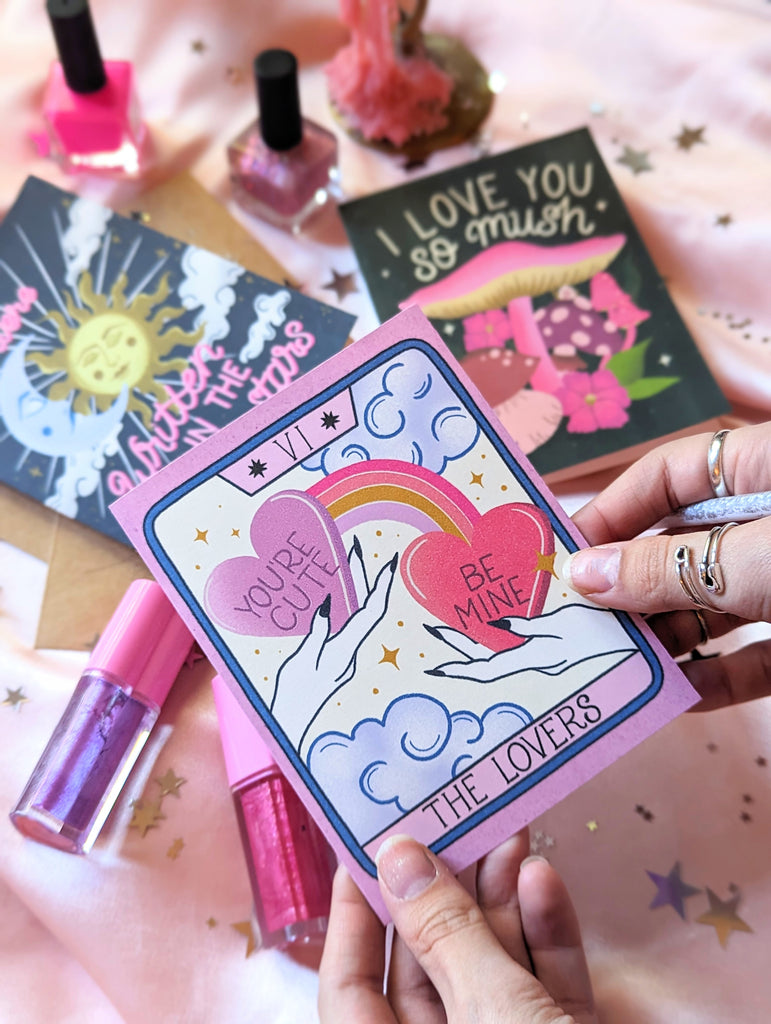 Three Valentine's Day cards sit on a pink background surrounded by star confetti and Trixie Cosmetics lip gloss. In the background one says "We Were Written in the Stars" in hand lettering with illustrations of a celestial sun and moon. There is another that says "I love you so mush" with whimsical mushrooms and fungi. In front someone holds up The Lovers Tarot Card, showing two hands with candy hearts, rainbows, and whimsical clouds all around.