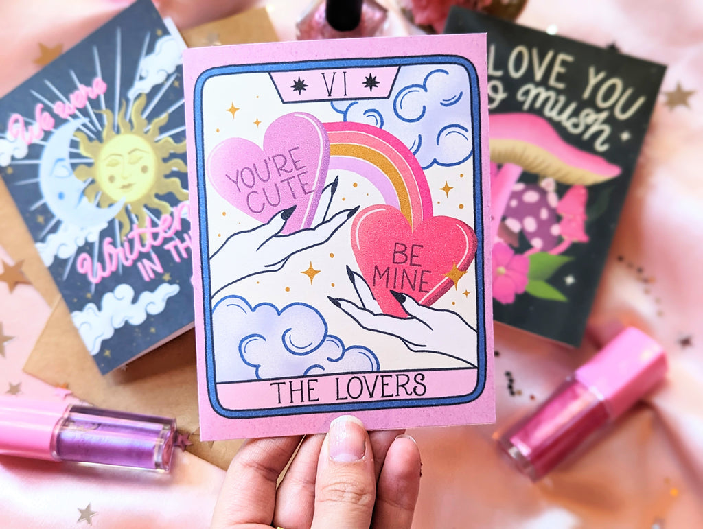 Three Valentine's Day cards are shown on a pink background with start confetti and Trixie Cosmetics lipgloss scattered around. One is being held up. The card features artwork inspired by The Lovers tarot card. There are two hands each holding a candy heart with hand lettering that says "You're Cute" and "Be Mine" A rainbow connects the hearts and in the background there are whimsical clouds and twinkling stars. A truly magical card to celebrate your love.