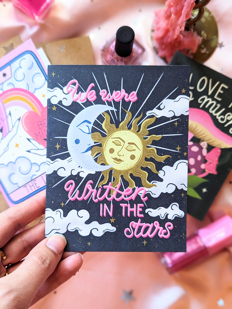 Three art prints are shown surrounded by a scattering of star confetti, candles, and makeup. One is behind held up, the art features hand lettering spelling out "We were written in the stars" accompanied by a beautiful celestial scene with a crescent moon and sun illuminating the night sky filled with whimsical clouds and twinkling stars.