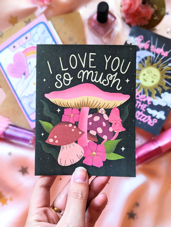 Three art prints are shown amongst scattered star confetti and glittery makeup. One print is being held up, it says "I Love you so mush" in a hand lettering script style. The illustration is of vibrant mushroom and fungi, with florals, twinkling stars, and an adorable little snail.