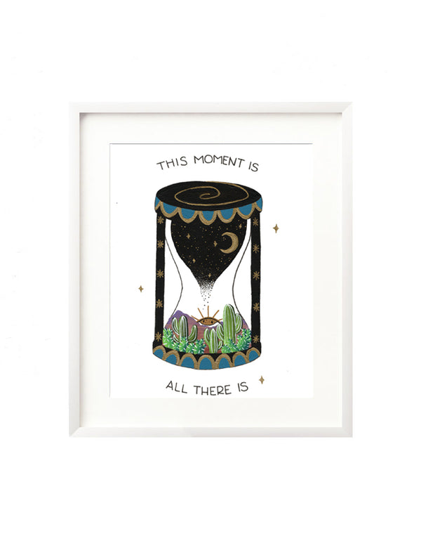 A framed art print - the illustration is of an hour glass with a scene of the dessert at sunset inside. There are lovely painted canyons, cacti, and the sun is an intuitive eye setting in the background. The hourglass stand contains a starry night sky, that drips into the bottom of the hourglass. Hand lettering spells out "This moment is all there is"