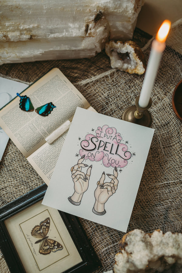 An art print sits atop an open book surrounded by witchy items such as crystals, preserved butterflies, and twinkling candles. The illustration is of two witchy hands adorned in celestial rings summoning a cloud of purple smoke. It is surrounded by twinkles and stars, and inside hand lettering spells out "I Put a Spell on You" inspired by Hocus Pocus, and perfect for Halloween.
