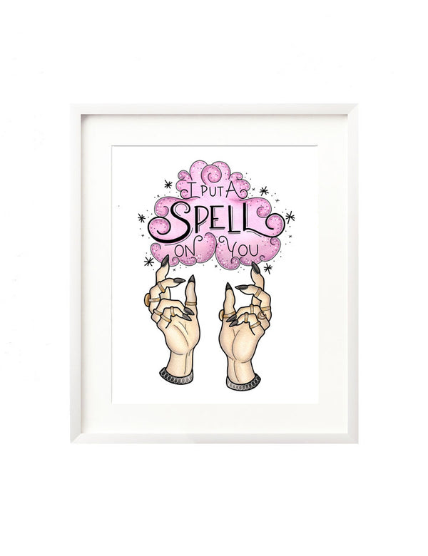 A framed art print - the illustration is of two witchy hands adorned with gold rings summoning a cloud of purple smoke. It is surrounded by twinkles and dots, and inside is the hand lettered message "I put a spell on you" inspired by the movie Hocus Pocus. A lovely addition to your Halloween decor. 